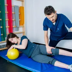 Fizjoterapeuta pomaga pacjentce w ćwiczeniach pnf. Kobieta leży na macie z żółtą piłką, a mężczyzna delikatnie naciska na jej nogę.