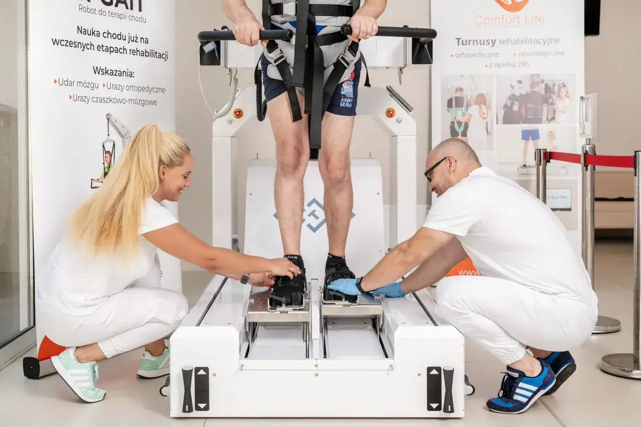 Rehabilitacja po złamaniu kości udowej: pacjent w specjalistycznym robocie do nauki chodu, asystują fizjoterapeuci.